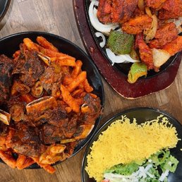 Three Indian dishes are displayed: a bowl of spicy curry with meat and vegetables, a sizzling platter of grilled meats with onions and peppers, and a side of yellow rice with salad.