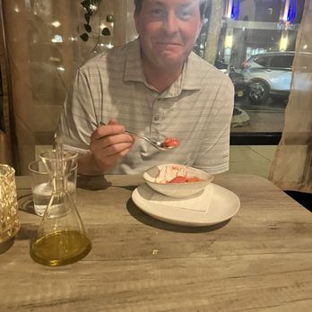 Ember & Cork - A man in a striped shirt is seated at a restaurant table, holding a spoon with a red-colored dish. The table has a glass of water, a small glass container, and a yellow oil bottle. - Seafood Near Me - Schenectady, New York