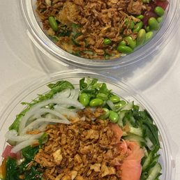 two bowls of food - Two clear plastic bowls hold Asian-style salads with shredded meat, green peas, lettuce, sliced onions, and various vegetables. The salads are topped with shredded meat and garnished with pickled ginger, sauces, and herbs. The presentation emphasizes fresh ingredients and colorful toppings, suitable for takeout or casual dining.