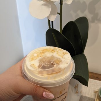 TP Tea - French brulee strawberry milk tea - A hand with manicured nails holds a strawberry milk tea in a clear plastic cup with a sealed lid, showing a creamy, pinkish beverage with some foam or toppings on top. The background features a potted orchid with white flowers and dark green leaves, placed on a table with a textured cloth. The setting appears to be indoors, possibly in a cafe or home environment. - Bubble Tea Near Me - Richardson, Texas