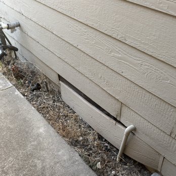 3 Generations Improvements - The before, with the siding starting to come loose. - The image depicts the side of a building with horizontal siding, where a section of siding is broken and displaced, revealing an opening to the foundation vent. The area around the vent has dirt, dried plants, and debris. A pipe runs vertically along the wall, and part of a bicycle is visible on the left side. The scene is outdoors, next to a concrete sidewalk. - Windows Installation Near Me - Folsom, California