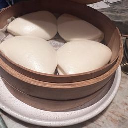 Four steamed buns with smooth, shiny surfaces are arranged inside a round wooden steamer basket. The basket is placed on a speckled white ceramic plate on a marble table, with some receipts or papers visible in the background. The buns appear freshly steamed and are likely part of Asian cuisine.