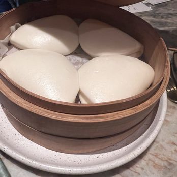The Monkey King - Four steamed buns with smooth, shiny surfaces are arranged inside a round wooden steamer basket. The basket is placed on a speckled white ceramic plate on a marble table, with some receipts or papers visible in the background. The buns appear freshly steamed and are likely part of Asian cuisine. - Chinese Near Me - Brooklyn, New York