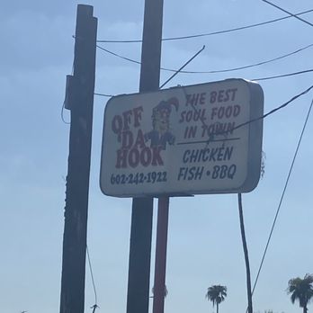 Stacy's Off Da Hook - The image shows an outdoor sign for Off Da Hook, a soul food restaurant, mounted on a pole with power lines overhead. The sign features the restaurant's name, phone number, and mentions menu items such as chicken, fish, and BBQ. There is a cartoon character on the sign wearing a jester hat. In the background, there are palm trees and a partly cloudy sky, indicating a warm climate. - Barbeque Near Me - Phoenix, Arizona
