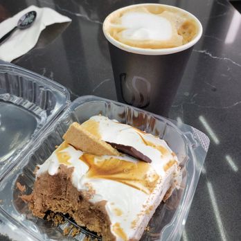 Sada Coffee - A disposable black coffee cup filled with a frothy coffee beverage is on a black marble table. Next to it, there is a plastic container with a partially eaten slice of cake topped with white icing, caramel drizzle, and a small cookie. A spoon and napkin are also visible on the table, and a person wearing a black jacket with white stripes is seated nearby. - Desserts Near Me - Palos Hills, Illinois