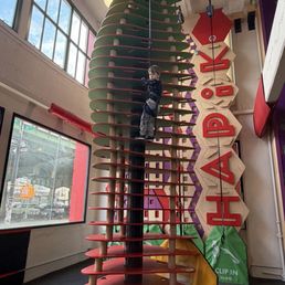 A boy is climbing a tall, spiral-shaped indoor play structure with horizontal steps, inside a play area with large windows and colorful decor.