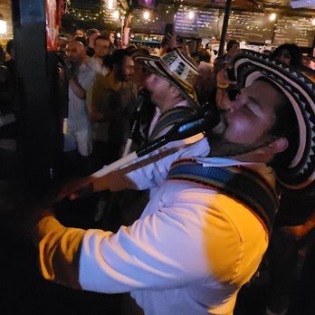 Terraza 7 - A man in a striped hat and white jacket is playing a keyboard at a crowded indoor venue with string lights and a bar in the background. - Bars Near Me - Elmhurst, New York