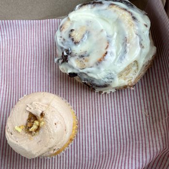 BATTER - Cinnamon roll and cornbread cup cake - A cinnamon roll with white icing and a cupcake with beige frosting and a small topping are placed on red and white striped paper inside a bakery box. - Desserts Near Me - Hammond, Louisiana