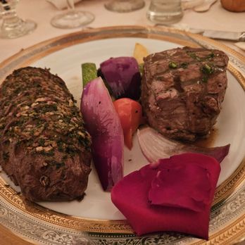 The Haley Mansion - A plate contains two portions of cooked meat, one seasoned with herbs and the other with a sauce. The meal is garnished with vegetables such as onion, tomato, eggplant, and zucchini, arranged around the meats. The plate has an ornate gold and white border, and the setting appears to be a formal dining environment with glasses and utensils visible in the background. - Party & Event Planning Near Me - Joliet, Illinois