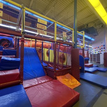 Off The Wall Gamezone - Tunnels - A spacious indoor playground with padded climbing structures, slides, and tunnels designed for children. The area includes colorful foam mats and safety signs on the upper railing indicating rules such as no pushing or throwing balls. The ceiling has bright yellow lighting, and the background shows shelves and colorful wall decorations. - Indoor Playcenter Near Me - Miami, Florida