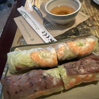 Pho Cafe - Combination Spring Rolls. - Two spring rolls, one with shrimp and lettuce and the other with meat, are served on a plate with a cup of tea in the background. - Vietnamese Near Me - Lafayette, Colorado