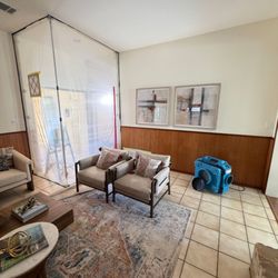 Twin Pillars Restoration - A furnished living room is partially sealed with clear plastic containment barriers while a blue air filtration machine opera...