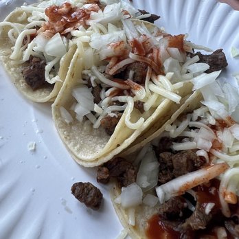La Costa - Carne Asada Tacos - Two carne asada tacos filled with grilled beef, topped with chopped onions, shredded cheese, and drizzled with sauce, served on a white disposable plate. The tacos are placed on a pinkish table surface, with some scattered meat pieces and onion bits around. The image captures a casual outdoor dining setting, typical of Mexican eateries offering tacos with simple toppings. - Mexican Near Me - Tracy, California