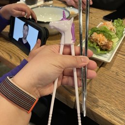 Pro and beginners chopsticks here! Unicorn shown in photo. Shark edition not shown. - A person holds a pair of unicorn-shaped chopsticks with a purple mane and a white horn, positioned over a wooden dining table. In the background, there are glasses of water, a plate with salad, and a person taking a photo with a smartphone. The setting appears to be a restaurant with a casual dining atmosphere.