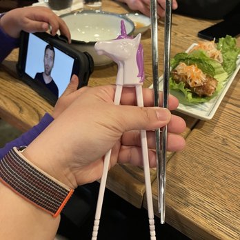 Pho Junkies - Pro and beginners chopsticks here! Unicorn shown in photo. Shark edition not shown. - A person holds a pair of unicorn-shaped chopsticks with a purple mane and a white horn, positioned over a wooden dining table. In the background, there are glasses of water, a plate with salad, and a person taking a photo with a smartphone. The setting appears to be a restaurant with a casual dining atmosphere. - Food Trucks Near Me