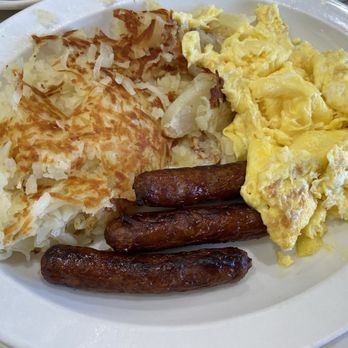 Alexander's Restaurant - Hash brown, sausage, scrambled eggs - A breakfast plate includes scrambled eggs, three cooked sausage links, hash browns, and mashed potatoes with gravy, all arranged on a white oval plate. The eggs are fluffy and yellow, the sausage links are browned, the hash browns are crispy, and the mashed potatoes are topped with gravy. The meal appears to be served in a casual dining setting. - Diners Near Me - Chicago, Illinois