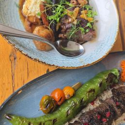 A plate of grilled meat with rice, topped with pomegranate seeds, accompanied by a roasted green chili and cherry tomatoes.