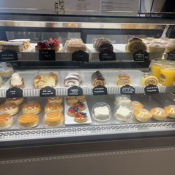 My Brigadeiro - The image shows a refrigerated display case filled with an assortment of desserts, cakes, and beverages. The top shelf features slices of cake, fruit tart, flan, and other pastries, each labeled with small black tags. The middle shelf contains cheesecakes, raspberry cream rolls, and chocolate rocamble, along with small fruit-topped desserts and cups of mango smoothies. The bottom shelf displays individual-sized puddings and custards. The display is well-organized, with clear labels and a variety of sweet treats suitable for a bakery or cafe setting. - Chocolatiers & Shops Near Me - Hanover, New Hampshire
