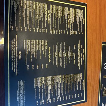 L’amour Restaurant - Menu - A menu printed on black paper with gold borders, listing various Indian, Nepalese, Pakistani, and Mediterranean dishes. It includes sections for soups, appetizers, house specials, and vegetarian options. The menu details prices and descriptions for each item, such as chicken tikka masala, naan breads, and vegetable dishes. It is placed on a wooden table, with part of another menu visible nearby. - Indian Near Me - Springfield, Massachusetts