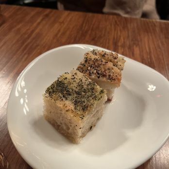 Citronnelle - Homemade focaccia - Two pieces of homemade focaccia bread, topped with herbs, spices, and seasonings, are placed on a white plate. The bread appears to be freshly baked with a golden crust and a soft interior. The plate is set on a wooden table, and the background shows a blurred view of the restaurant interior with a person seated nearby. - French Near Me - Lancaster, Pennsylvania