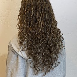 The color and cut both were perfection. - A person with long, naturally curly hair viewed from behind, wearing a gray hoodie, standing against a textured white wall.