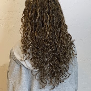 Roots Salon - The color and cut both were perfection. - A person with long, naturally curly hair viewed from behind, wearing a gray hoodie, standing against a textured white wall. - Hair Salons Near Me - Albuquerque, New Mexico