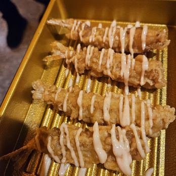 Y Space BBQ - Four fried fish skewers are arranged on a gold-colored tray, each topped with a zigzag drizzle of mayonnaise. The skewers are golden-brown and appear crispy. The tray has a textured surface, and part of a person's leg and shoe are visible in the background, indicating an outdoor or casual dining setting. - Barbeque Near Me - Flushing, New York