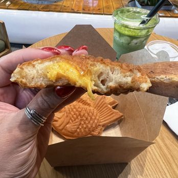 Brucha - A person holds a partially eaten pastry with yellow filling, showing its soft interior, over a fish-shaped pastry on a wooden table. In the background, there is a green drink with a straw, and the setting appears to be a cafe with potted plants and wooden furniture. - Gelato Near Me - Tucson, Arizona