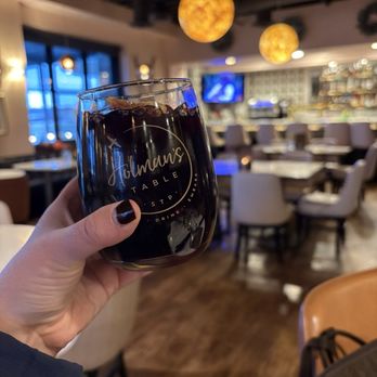 Holman's Table - Sangria - A person is holding a glass of dark soda with ice in a restaurant setting. The glass has the logo 'Holman's Table' on it. The background features a dining area with tables, chairs, a bar, and hanging spherical lights. The restaurant has a modern interior with large windows and ambient lighting. - Bars Near Me - Saint Paul, Minnesota