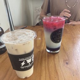Chai Tea Latte - Two drinks are placed on a wooden table at Fluid Coffee Roasters, one is a creamy iced beverage and the other is a layered drink with red, white, and dark layers.