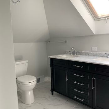 360 Improvements - a bathroom with a skylight - The bathroom has a sloped ceiling with a large skylight window allowing natural light. It contains a white toilet positioned near the wall and a black vanity with a marble countertop and dual sinks. The floor is tiled with white marble-patterned tiles, and a towel bar is mounted on the wall above the vanity. An electrical outlet is visible on the wall near the countertop. - General Contractors Near Me - Plano, Illinois