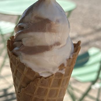 Pink Pony Soft Serve - A soft serve ice cream cone with chocolate and vanilla swirls is held outdoors, with a blurred background of chairs and a table. - Ice Cream & Frozen Yogurt Near Me - Albuquerque, New Mexico