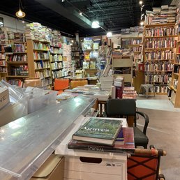 Back - The image shows the interior of a bookstore with numerous wooden bookshelves filled with books of various sizes and colors. A table in the foreground holds stacks of books and papers, with some chairs and boxes around it. The ceiling is black with industrial lighting, and the space appears densely packed with books and reading materials, creating a cluttered but organized environment.