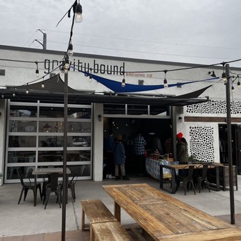 Gravity Bound Brewing - The image depicts the outdoor patio of Gravity Bound Brewing in Albuquerque, NM, featuring string lights hanging above the seating area. There are wooden and metal tables and chairs, with some people standing near the entrance. The building has a white exterior with large garage-style windows and a sign reading 'gravity bound brewing co.' The scene suggests a casual, outdoor gathering space for patrons of the brewery. - Brewpubs Near Me - Albuquerque, New Mexico