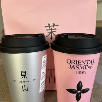 Molly Tea - Two disposable coffee cups from Molly Tea are positioned in front of a pink Molly Tea shopping bag. The pink cup is labeled 'Oriental Jasmine' with a black floral graphic, while the silver cup is labeled 'Jianshan' with Chinese characters and additional product information. Both cups have black lids and are set on a textured surface. - Bubble Tea Near Me - Markham, Ontario