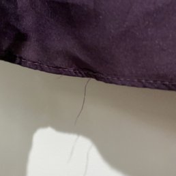 Threads hanging loose - The image depicts a close-up view of purple fabric, likely part of a garment, with visible loose threads hanging from the hem area. The fabric appears to be slightly wrinkled, and the loose threads suggest it may require sewing or alteration. The background is a plain, light-colored surface.