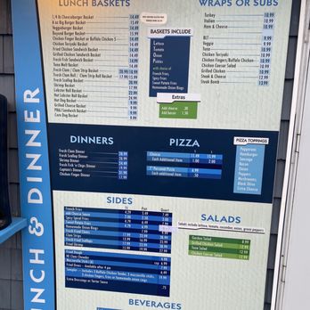 Oceanside Store - Lunch and dinner menu - The menu board features sections for lunch baskets, wraps or subs, dinners, pizzas, sides, salads, and beverages. It includes prices for various food items such as burger baskets, chicken, seafood, and pizza toppings. The board also lists salad options and drink choices, with additional charges for extras like cheese and bacon. The menu indicates availability times and notes that pizza is not served after 11 am. The layout is organized with color-coded sections and pricing details. - Convenience Stores Near Me - York, Maine
