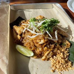Pad Thai - A dish of Pad Thai is presented on a tray lined with parchment paper. It includes stir-fried rice noodles with chicken, topped with bean sprouts and cilantro. A lime wedge and crushed peanuts are placed on the side. The tray is set on a wooden table, with a glass of water and a white plate visible in the background.