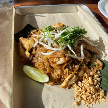 Charm Thai Eatery - Pad Thai - A dish of Pad Thai is presented on a tray lined with parchment paper. It includes stir-fried rice noodles with chicken, topped with bean sprouts and cilantro. A lime wedge and crushed peanuts are placed on the side. The tray is set on a wooden table, with a glass of water and a white plate visible in the background. - Thai Near Me - Westminster, Colorado