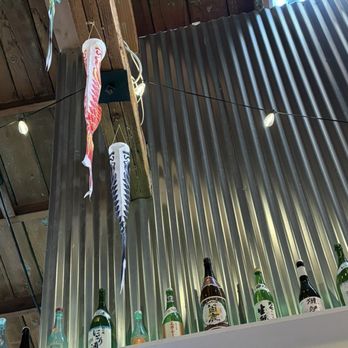 Kinda Izakaya - interior - Fish-shaped windsocks, known as koinobori, hang from a wooden beam in a restaurant interior. The background features a corrugated metal wall and a shelf with various bottles of sake. String lights are also visible, adding to the decor. - Tapas/Small Plates Near Me - Berkeley, California