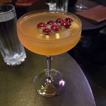 Rouge 41 - Carda nom-nom - A cocktail with a pinkish-orange hue is presented in a coupe glass, garnished with red berries floating on the surface. The drink is placed on a dark, textured table, with two tall glasses of water and a leather-bound menu visible in the background. The setting appears to be a bar or restaurant with dim lighting and a social atmosphere. - Speakeasies Near Me - Chaska, Minnesota