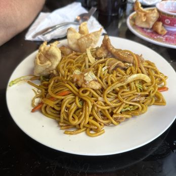 Bao Bao Chinese Bistro - 10. Meat Lo Mein Dinner - A white plate contains meat lo mein noodles mixed with vegetables such as cabbage and carrots, topped with fried wontons. The plate is on a dark table with a fork, napkin, and other dishes and drinks visible in the background, indicating a restaurant setting. - Chinese Near Me - Springfield, Missouri