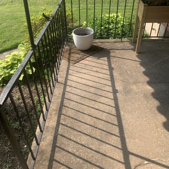 A Hoffman Awning - The image shows a small outdoor balcony with a black metal railing, casting diagonal shadows on the concrete floor. There is a white round planter with soil placed on a small mat, and a wooden plant stand with a rectangular planter on top. The background includes a grassy yard with plants and a brick building wall. - Awnings Near Me - Baltimore, Maryland
