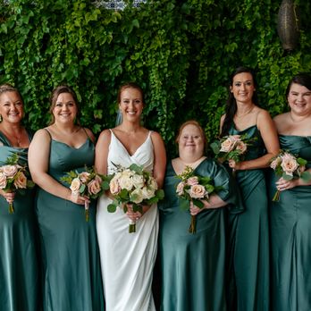 Daydream Events - A bride and seven bridesmaids stand in a row holding bouquets, with a lush green leafy wall behind them, during a wedding celebration. - Wedding Planning Near Me