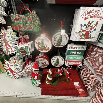At Home - A display of Christmas decorations such as ornaments, figurines, and signs with holiday messages, arranged on a store shelf. - Furniture Stores Near Me - Merrillville, Indiana