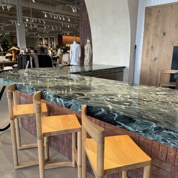 A-OK Cafe - Bar seating - A marble bar counter with a dark green and white veined surface is positioned in a retail store environment. Three wooden chairs with light wood finish are placed along the counter. In the background, clothing displays and mannequins dressed in white and beige garments are visible, with store lighting and ceiling fixtures overhead. The setting appears to be a clothing or lifestyle retail store with modern interior design elements. - Coffee & Tea Near Me - Vaughan, Ontario