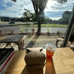 Cold brew and cinnamon roll to go! - Inside a cafe, a wooden table holds food containers and iced tea, with a large window revealing outdoor seating, a tree, and a partly cloudy sky.