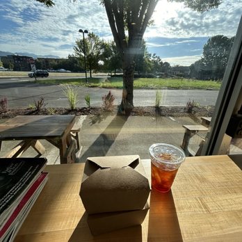Southpine Bakery - Cold brew and cinnamon roll to go! - Inside a cafe, a wooden table holds food containers and iced tea, with a large window revealing outdoor seating, a tree, and a partly cloudy sky. - Coffee & Tea Near Me - Eugene, Oregon