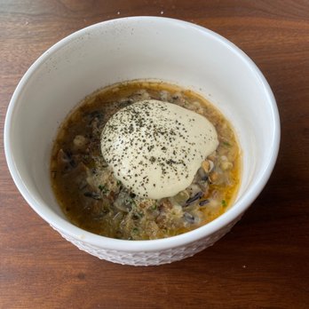 Canoe - A white bowl contains a soup with chopped vegetables and herbs, topped with a dollop of cream and black pepper, placed on a wooden surface. - Canadian (New) Near Me - Toronto, Ontario