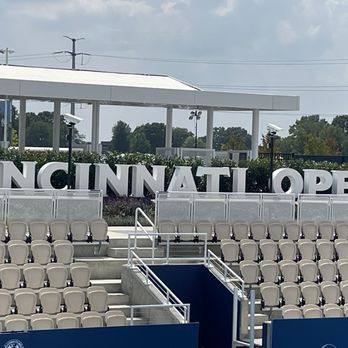 HangOverEasy - Cincinnati Open not far from this joynt. - The image shows an outdoor tennis stadium with multiple rows of beige seats, some stairs, and railings. Large white letters spell out 'CINCINNATI OPEN' on a hedge behind the seating area. The sky is partly cloudy, and there are trees and utility poles in the background. A person wearing a hat is visible near the left side of the seating area. - Breakfast & Brunch Near Me - Cincinnati, Ohio